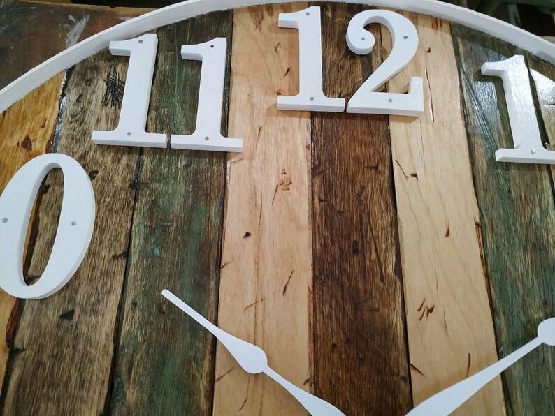 May include: A white clock face with black Roman numeral hour markers on a rustic wooden background. The clock face is made of wood planks in various shades of brown, green, and beige. The clock hands are white and point to the numbers 11 and 12.