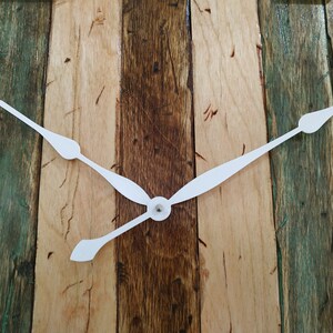 May include: White clock hands on a wooden background. The wood is a mix of light and dark brown with green accents.