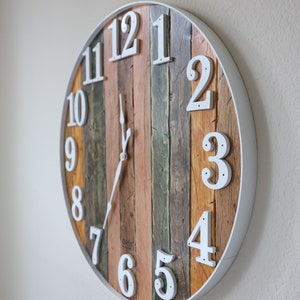 May include: A round, white-framed clock with wooden planks for the face. The numbers are white and the hands are black. The clock is hanging on a white wall.