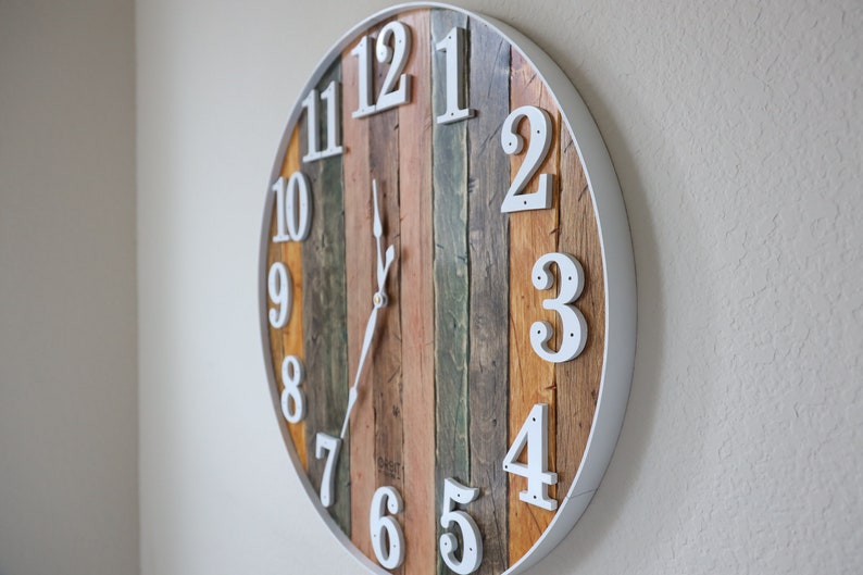 May include: A round, white-framed clock with wooden planks for the face. The numbers are white and the hands are black. The clock is hanging on a white wall.