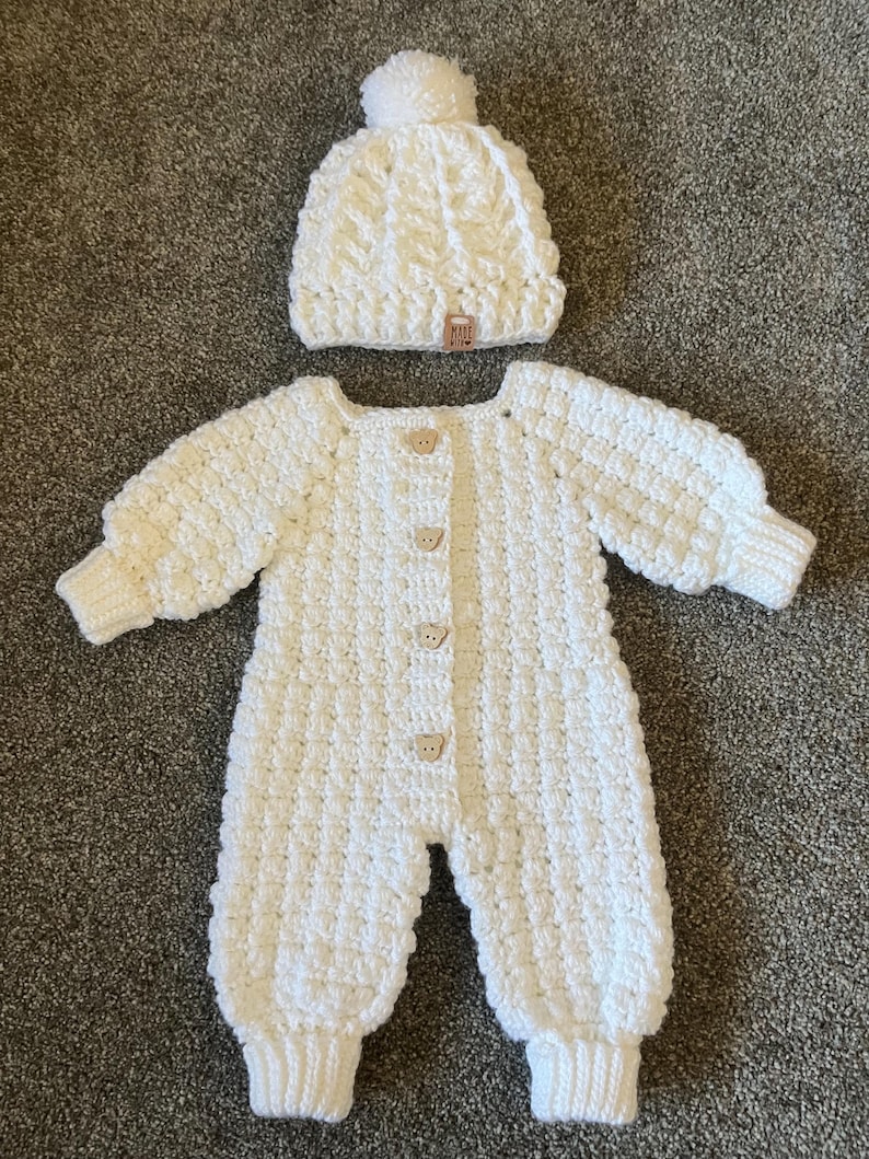 Puede incluir: Un pelele de manga larga de ganchillo blanco con patr&oacute;n texturizado, cuatro botones de color marr&oacute;n claro con forma de oso en la parte delantera y pu&ntilde;os acanalados. Incluye un gorro de ganchillo blanco a juego con pomp&oacute;n y etiqueta MADE WITH.
