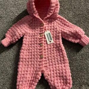 May include: A light pink crocheted hooded jumpsuit featuring a textured bobble stitch pattern, long sleeves with ribbed cuffs, and four small bear-shaped buttons down the front. A white rectangular product tag is attached to the top button.