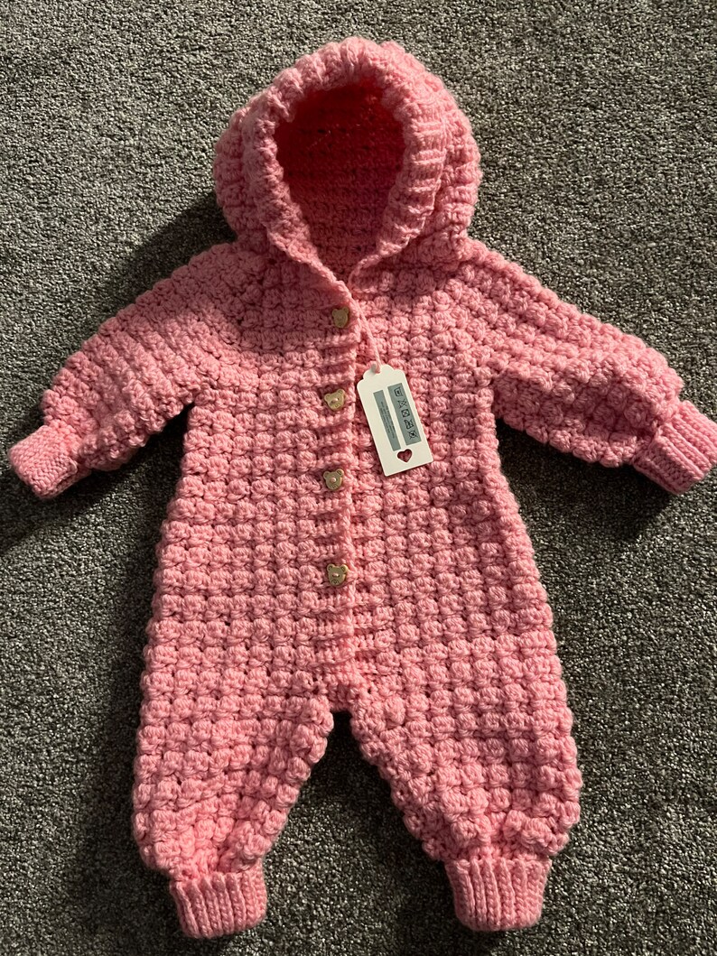 May include: A light pink crocheted hooded jumpsuit featuring a textured bobble stitch pattern, long sleeves with ribbed cuffs, and four small bear-shaped buttons down the front. A white rectangular product tag is attached to the top button.