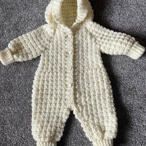 May include: A cream-colored, long-sleeved, hooded crochet romper featuring a textured waffle-like stitch pattern. The garment includes ribbed cuffs at the wrists and ankles, and is fastened down the front with four small, light-colored bear-shaped buttons.