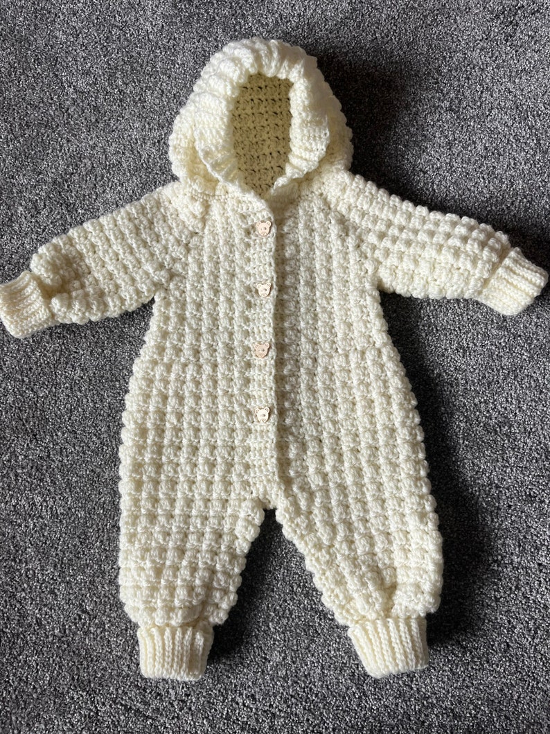 May include: A cream-colored, long-sleeved, hooded crochet romper featuring a textured waffle-like stitch pattern. The garment includes ribbed cuffs at the wrists and ankles, and is fastened down the front with four small, light-colored bear-shaped buttons.