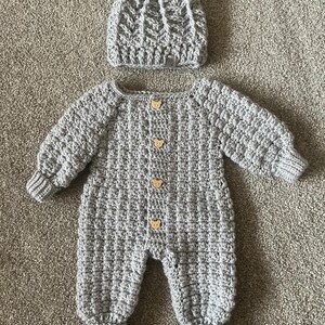 Puede incluir: Un pelele de manga larga tejido a ganchillo en color gris claro con un patr&oacute;n de puntos texturizados y cuatro peque&ntilde;os botones de madera con forma de oso en la parte delantera, junto a un gorro de ganchillo gris claro a juego con un pomp&oacute;n.