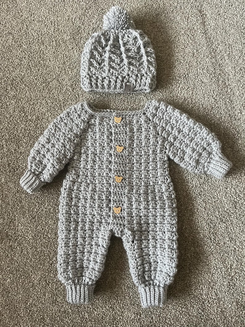Puede incluir: Un pelele de manga larga tejido a ganchillo en color gris claro con un patr&oacute;n de puntos texturizados y cuatro peque&ntilde;os botones de madera con forma de oso en la parte delantera, junto a un gorro de ganchillo gris claro a juego con un pomp&oacute;n.