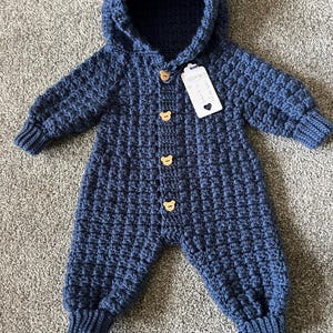 May include: A dark blue crocheted hooded jumpsuit featuring a textured waffle stitch pattern, long sleeves with ribbed cuffs, and four light brown wooden buttons shaped like bear heads. A white rectangular tag is attached to the top button.