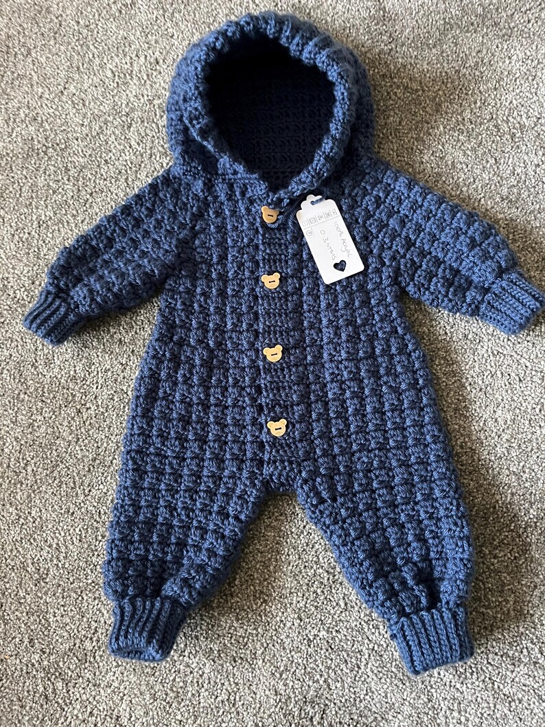 May include: A dark blue crocheted hooded jumpsuit featuring a textured waffle stitch pattern, long sleeves with ribbed cuffs, and four light brown wooden buttons shaped like bear heads. A white rectangular tag is attached to the top button.