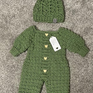Puede incluir: Un pelele de manga larga tejido en crochet color verde salvia con cuatro botones claros en forma de oso y una etiqueta rectangular blanca, junto a un gorro de crochet a juego en verde salvia con un pomp&oacute;n de piel sint&eacute;tica oscura.
