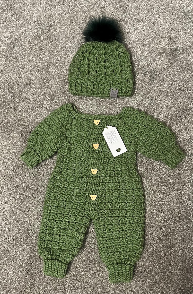 Puede incluir: Un pelele de manga larga tejido en crochet color verde salvia con cuatro botones claros en forma de oso y una etiqueta rectangular blanca, junto a un gorro de crochet a juego en verde salvia con un pomp&oacute;n de piel sint&eacute;tica oscura.