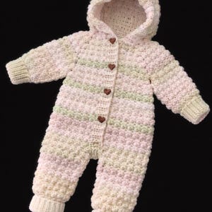 May include: A long-sleeved, hooded crochet romper featuring a textured bobble stitch pattern in horizontal stripes of pale pink, soft green, and cream. The front opening is secured with four small, dark brown, heart-shaped wooden buttons.