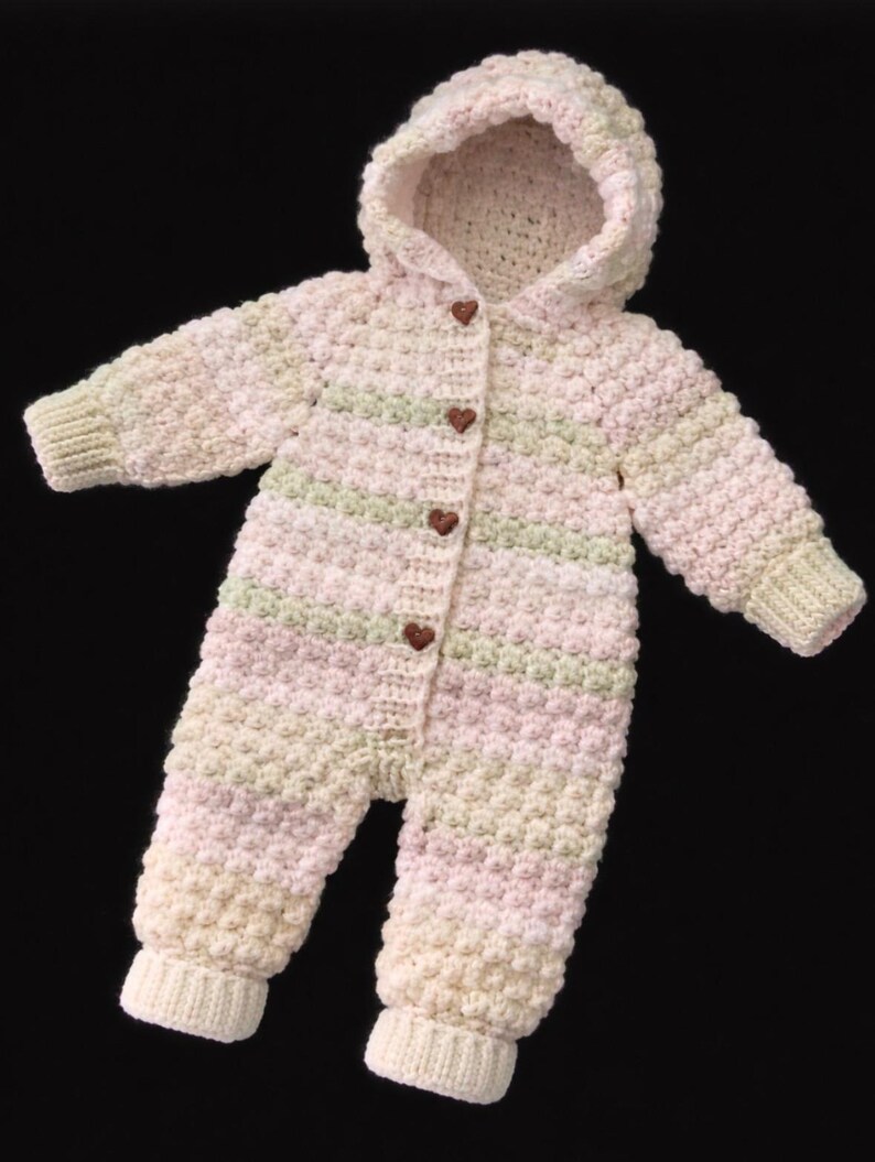 May include: A long-sleeved, hooded crochet romper featuring a textured bobble stitch pattern in horizontal stripes of pale pink, soft green, and cream. The front opening is secured with four small, dark brown, heart-shaped wooden buttons.