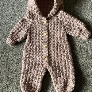 May include: A dusty rose crochet hooded jumpsuit featuring a textured bobble stitch pattern throughout. The garment includes long sleeves with ribbed cuffs, ribbed leg openings, and four light-colored wooden buttons shaped like bear faces.