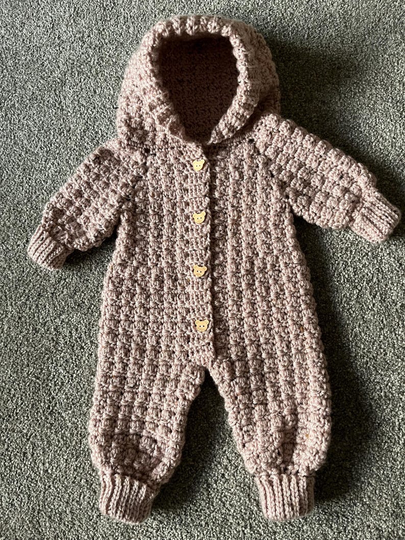 May include: A dusty rose crochet hooded jumpsuit featuring a textured bobble stitch pattern throughout. The garment includes long sleeves with ribbed cuffs, ribbed leg openings, and four light-colored wooden buttons shaped like bear faces.