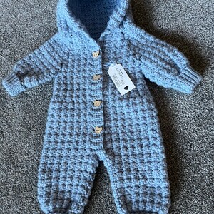 May include: A light blue hooded crochet jumpsuit featuring a textured waffle stitch pattern, long sleeves with ribbed cuffs, and four wooden buttons shaped like bear faces down the front. A white rectangular product tag is attached to the chest.