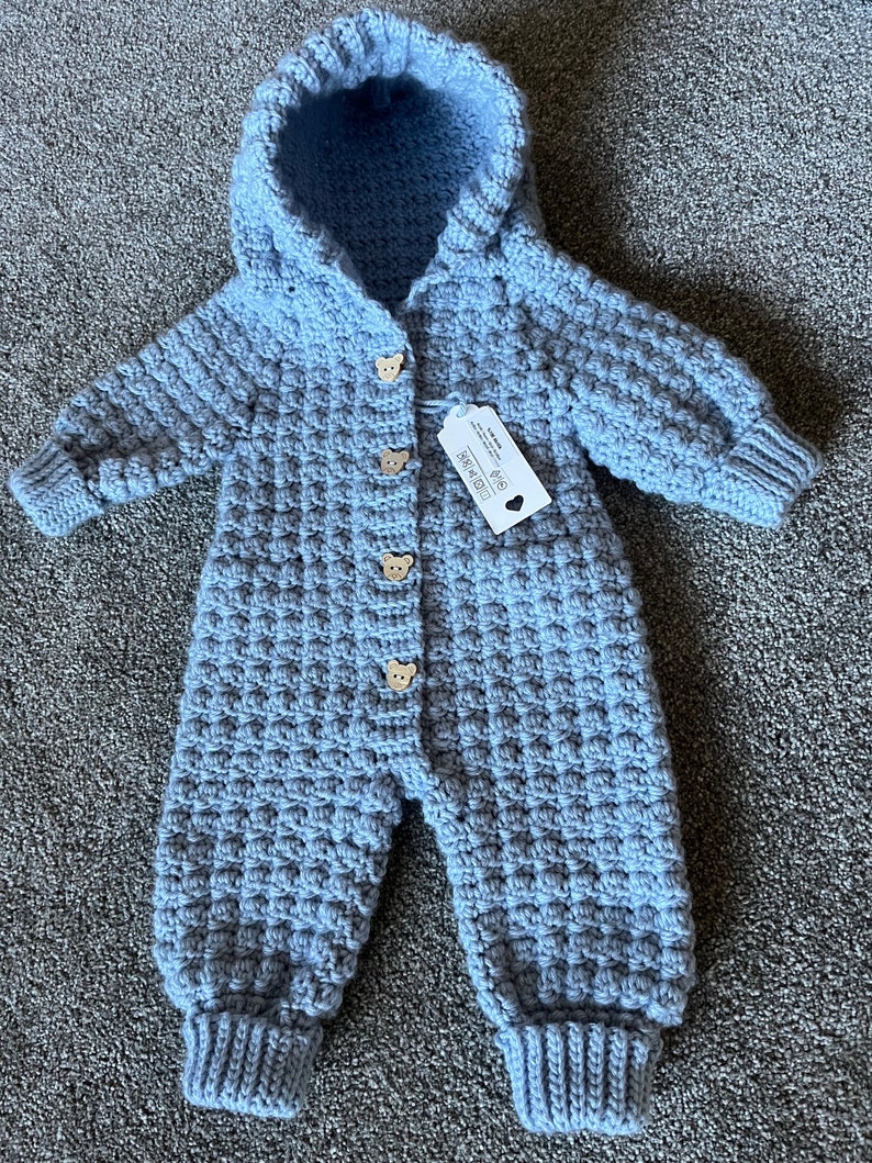 May include: A light blue hooded crochet jumpsuit featuring a textured waffle stitch pattern, long sleeves with ribbed cuffs, and four wooden buttons shaped like bear faces down the front. A white rectangular product tag is attached to the chest.