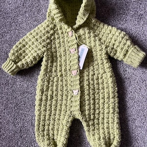 May include: A light green, long-sleeved, hooded crochet romper featuring a textured bobble stitch pattern. The garment includes four small, light-colored bear-shaped buttons down the front center and a white rectangular product tag attached.