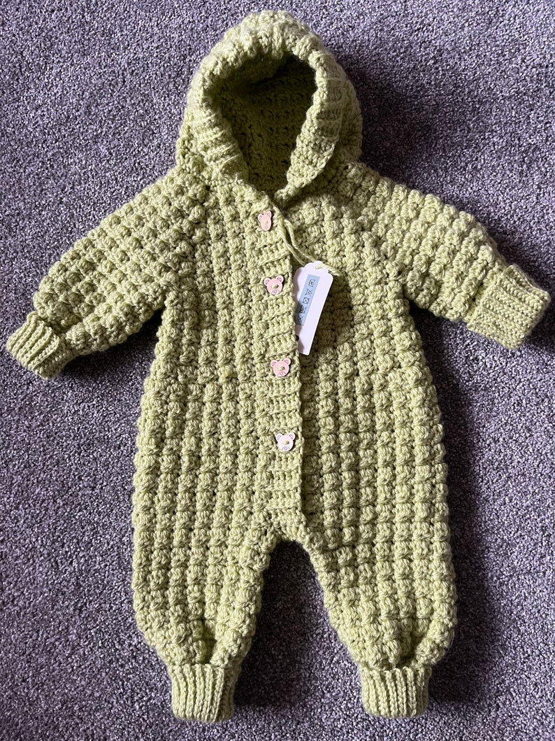May include: A light green, long-sleeved, hooded crochet romper featuring a textured bobble stitch pattern. The garment includes four small, light-colored bear-shaped buttons down the front center and a white rectangular product tag attached.