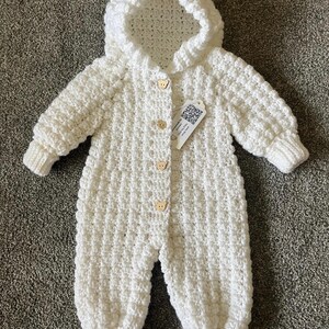 May include: A white crocheted long-sleeve jumpsuit with a hood, featuring a textured waffle-like stitch pattern and four light brown wooden buttons shaped like bear heads down the front center. A small white tag with a QR code is attached.