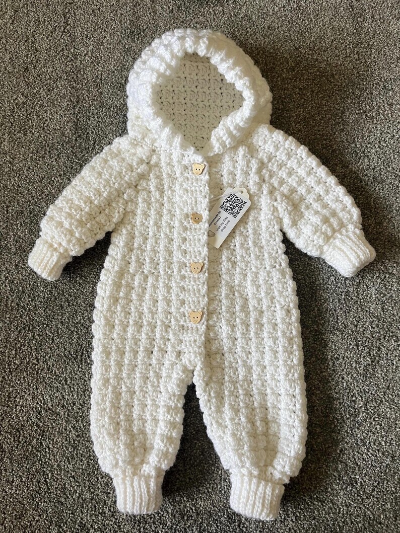 May include: A white crocheted long-sleeve jumpsuit with a hood, featuring a textured waffle-like stitch pattern and four light brown wooden buttons shaped like bear heads down the front center. A small white tag with a QR code is attached.