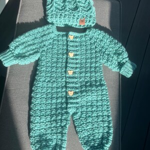 Puede incluir: Un pelele de ganchillo de manga larga en color verde azulado con un gorro de pomp&oacute;n a juego. El pelele presenta un patr&oacute;n de punto de gofre texturizado, pu&ntilde;os acanalados en mu&ntilde;ecas y tobillos, y cuatro botones de madera clara con forma de oso.