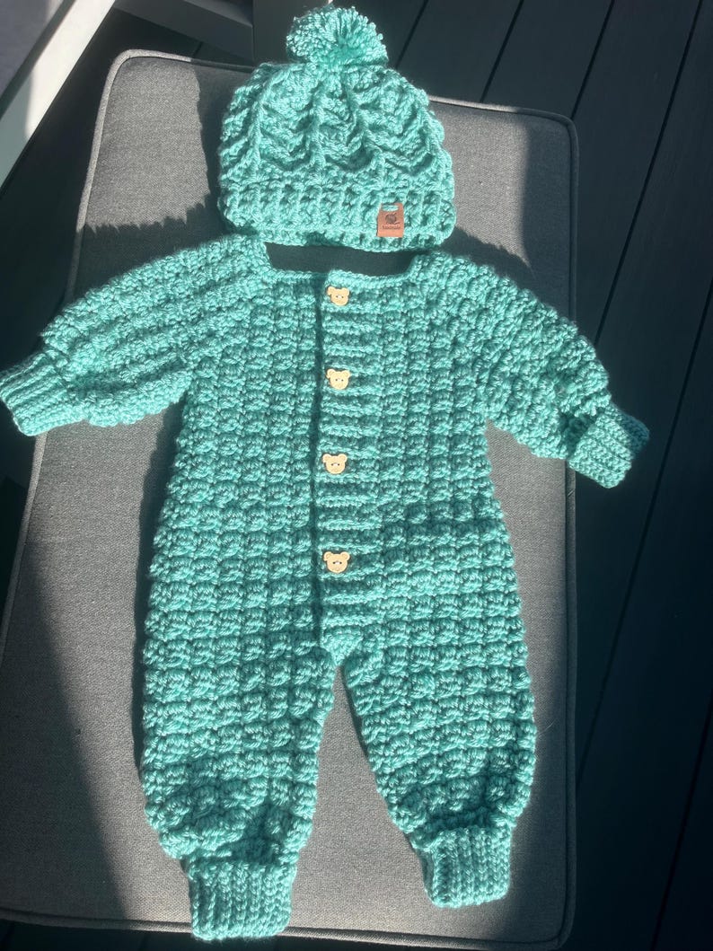 Puede incluir: Un pelele de ganchillo de manga larga en color verde azulado con un gorro de pomp&oacute;n a juego. El pelele presenta un patr&oacute;n de punto de gofre texturizado, pu&ntilde;os acanalados en mu&ntilde;ecas y tobillos, y cuatro botones de madera clara con forma de oso.