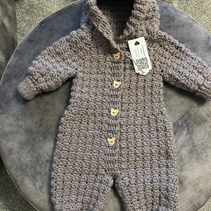 May include: A taupe-colored, long-sleeved, hooded crochet jumpsuit featuring a textured stitch pattern. The garment is fastened down the front with four light-colored wooden buttons shaped like bear faces and includes a white rectangular tag.