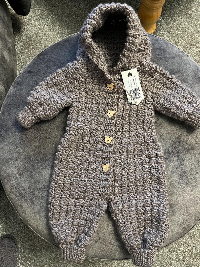 May include: A taupe-colored, long-sleeved, hooded crochet jumpsuit featuring a textured stitch pattern. The garment is fastened down the front with four light-colored wooden buttons shaped like bear faces and includes a white rectangular tag.