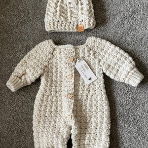 Puede incluir: Un pelele de beb&eacute; de ganchillo en color crema con mangas largas y pu&ntilde;os acanalados, que presenta cinco peque&ntilde;os botones de madera con forma de oso en la parte delantera. Incluye un gorro de ganchillo a juego con un pomp&oacute;n y una etiqueta Made With Love.