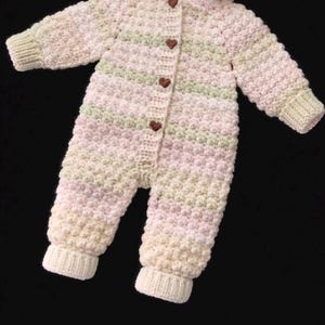 Puede incluir: Un pelele de ganchillo de manga larga con un patr&oacute;n de puntos texturizados en rayas horizontales de color rosa p&aacute;lido, verde suave y crema. La prenda incluye una tapeta frontal con cuatro peque&ntilde;os botones marrones en forma de coraz&oacute;n.
