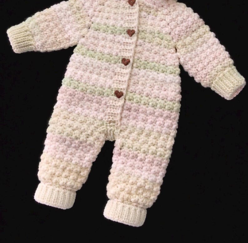 Puede incluir: Un pelele de ganchillo de manga larga con un patr&oacute;n de puntos texturizados en rayas horizontales de color rosa p&aacute;lido, verde suave y crema. La prenda incluye una tapeta frontal con cuatro peque&ntilde;os botones marrones en forma de coraz&oacute;n.