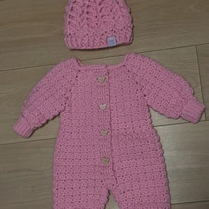 Puede incluir: Un pelele de manga larga tejido a ganchillo en color rosa claro con cuatro peque&ntilde;os botones de madera en forma de oso en la parte delantera y pu&ntilde;os acanalados. Incluye un gorro de ganchillo a juego con un pomp&oacute;n suave y una etiqueta blanca.