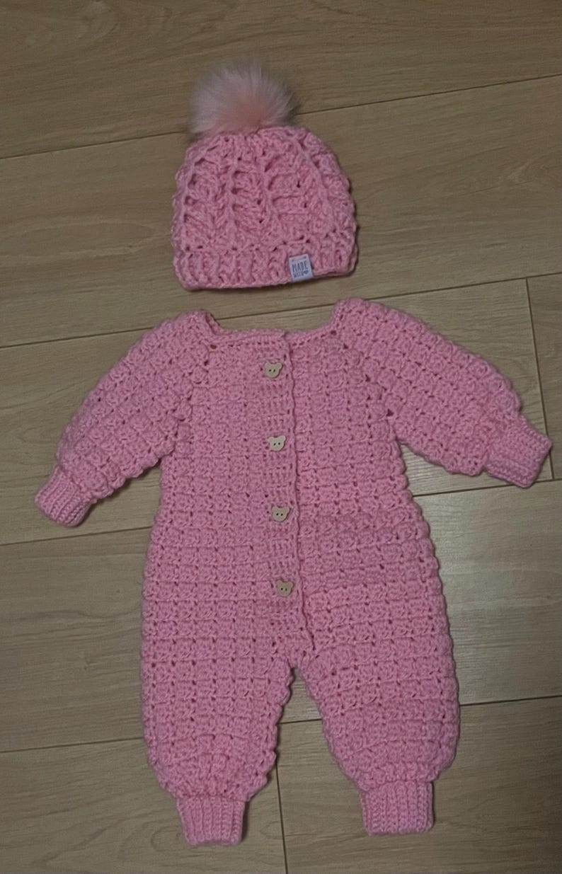 Puede incluir: Un pelele de manga larga tejido a ganchillo en color rosa claro con cuatro peque&ntilde;os botones de madera en forma de oso en la parte delantera y pu&ntilde;os acanalados. Incluye un gorro de ganchillo a juego con un pomp&oacute;n suave y una etiqueta blanca.