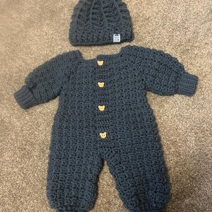 Puede incluir: Un pelele de manga larga de ganchillo en color gris oscuro con cuatro botones de color marr&oacute;n claro en forma de oso en la parte delantera y un gorro de ganchillo a juego con un pomp&oacute;n. El gorro tiene una peque&ntilde;a etiqueta blanca Made With Love.