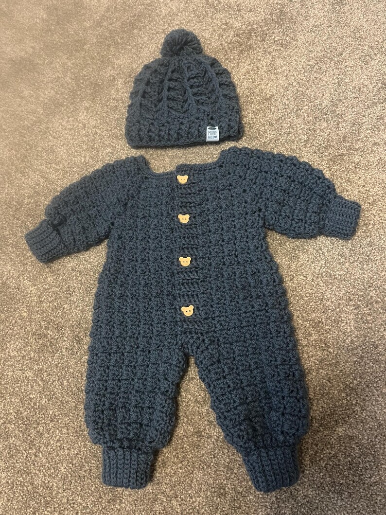 Puede incluir: Un pelele de manga larga de ganchillo en color gris oscuro con cuatro botones de color marr&oacute;n claro en forma de oso en la parte delantera y un gorro de ganchillo a juego con un pomp&oacute;n. El gorro tiene una peque&ntilde;a etiqueta blanca Made With Love.