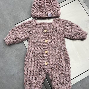 Puede incluir: Un mameluco de manga larga tejido a ganchillo en color rosa polvoriento y un gorro a juego con pomp&oacute;n. El mameluco presenta cuatro botones claros con forma de oso en la parte delantera. El hilo tiene una textura moteada con manchas oscuras.