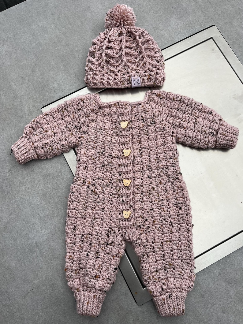 Puede incluir: Un mameluco de manga larga tejido a ganchillo en color rosa polvoriento y un gorro a juego con pomp&oacute;n. El mameluco presenta cuatro botones claros con forma de oso en la parte delantera. El hilo tiene una textura moteada con manchas oscuras.