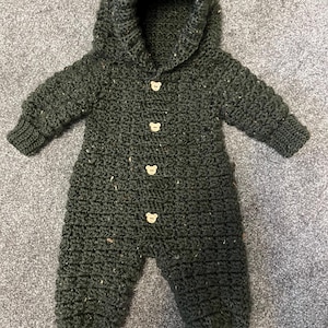 May include: A dark forest green crocheted one-piece jumpsuit with a hood, long sleeves, and ribbed cuffs at the wrists and ankles. The garment features four light-colored wooden buttons shaped like bear faces down the center front.