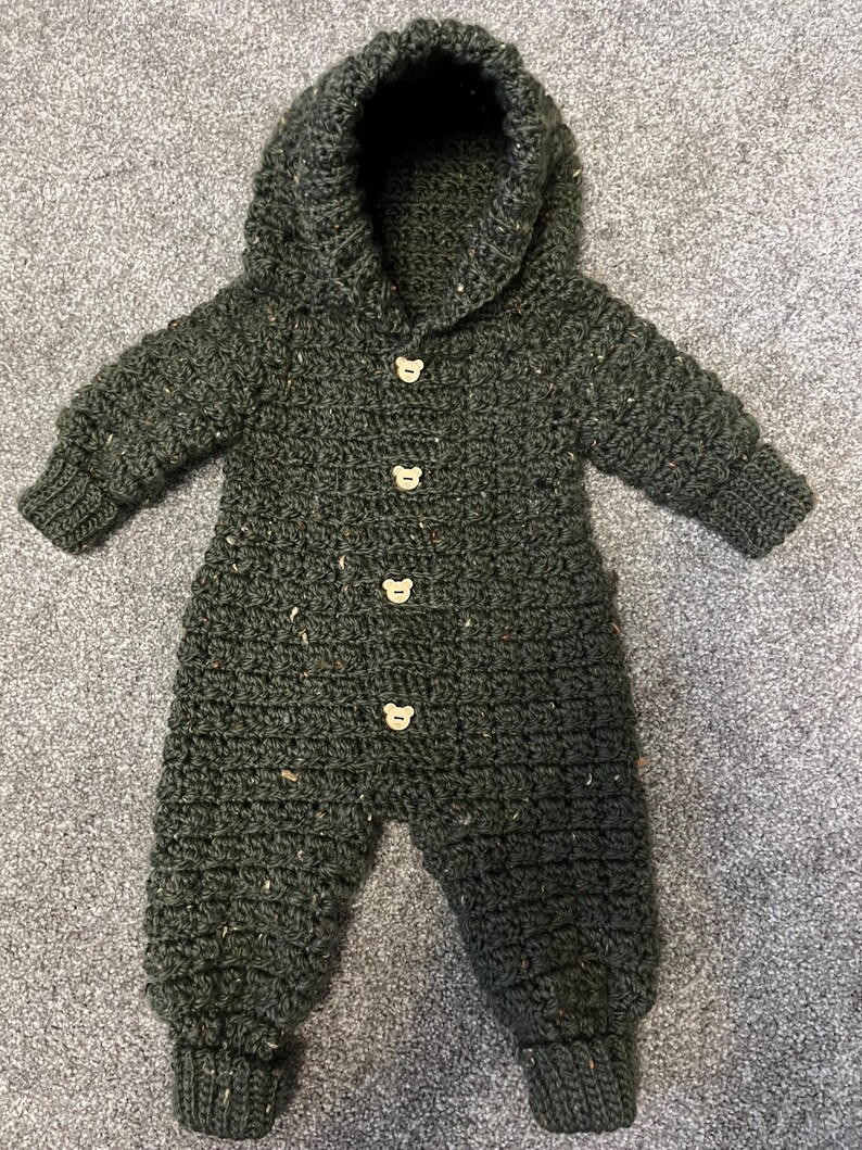 May include: A dark forest green crocheted one-piece jumpsuit with a hood, long sleeves, and ribbed cuffs at the wrists and ankles. The garment features four light-colored wooden buttons shaped like bear faces down the center front.