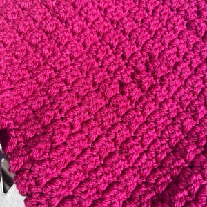 May include: A vibrant magenta crochet fabric featuring a dense, repeating textured stitch pattern. The yarn is worked in horizontal rows, creating a thick, raised surface with a consistent, uniform appearance throughout the entire visible textile.
