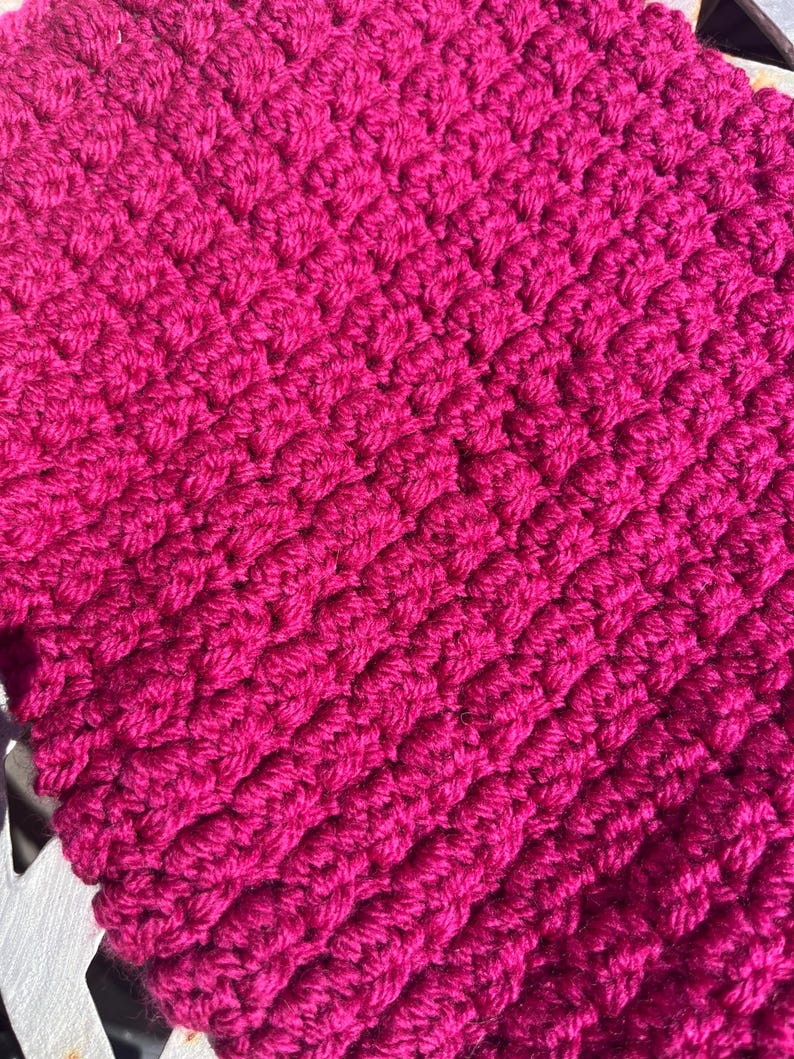 May include: A vibrant magenta crochet fabric featuring a dense, repeating textured stitch pattern. The yarn is worked in horizontal rows, creating a thick, raised surface with a consistent, uniform appearance throughout the entire visible textile.