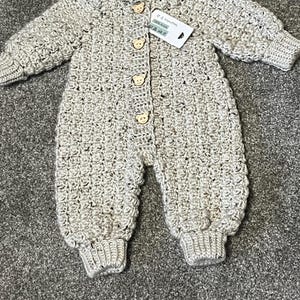 Puede incluir: Un pelele de manga larga tejido en crochet de color beige con un patr&oacute;n texturizado, pu&ntilde;os acanalados y cuatro botones de madera clara con forma de cara de oso en el centro. Una etiqueta blanca en el cuello muestra el texto 0-3 MONTHS.