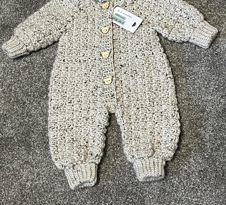 Puede incluir: Un pelele de manga larga tejido en crochet de color beige con un patr&oacute;n texturizado, pu&ntilde;os acanalados y cuatro botones de madera clara con forma de cara de oso en el centro. Una etiqueta blanca en el cuello muestra el texto 0-3 MONTHS.
