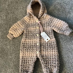May include: A long-sleeved, hooded crochet romper featuring a variegated pattern of beige, brown, and cream yarn. The garment includes five small, light-colored bear-shaped buttons down the front and a white rectangular tag attached with a pink string.