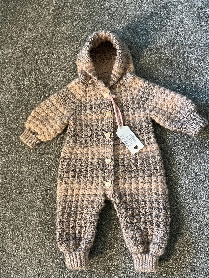 May include: A long-sleeved, hooded crochet romper featuring a variegated pattern of beige, brown, and cream yarn. The garment includes five small, light-colored bear-shaped buttons down the front and a white rectangular tag attached with a pink string.