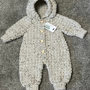 May include: A beige crochet hooded romper featuring long sleeves, ribbed cuffs, and four light brown bear-shaped wooden buttons down the front. A white rectangular tag is attached, displaying the text 0-3 months and 100% Acrylic.