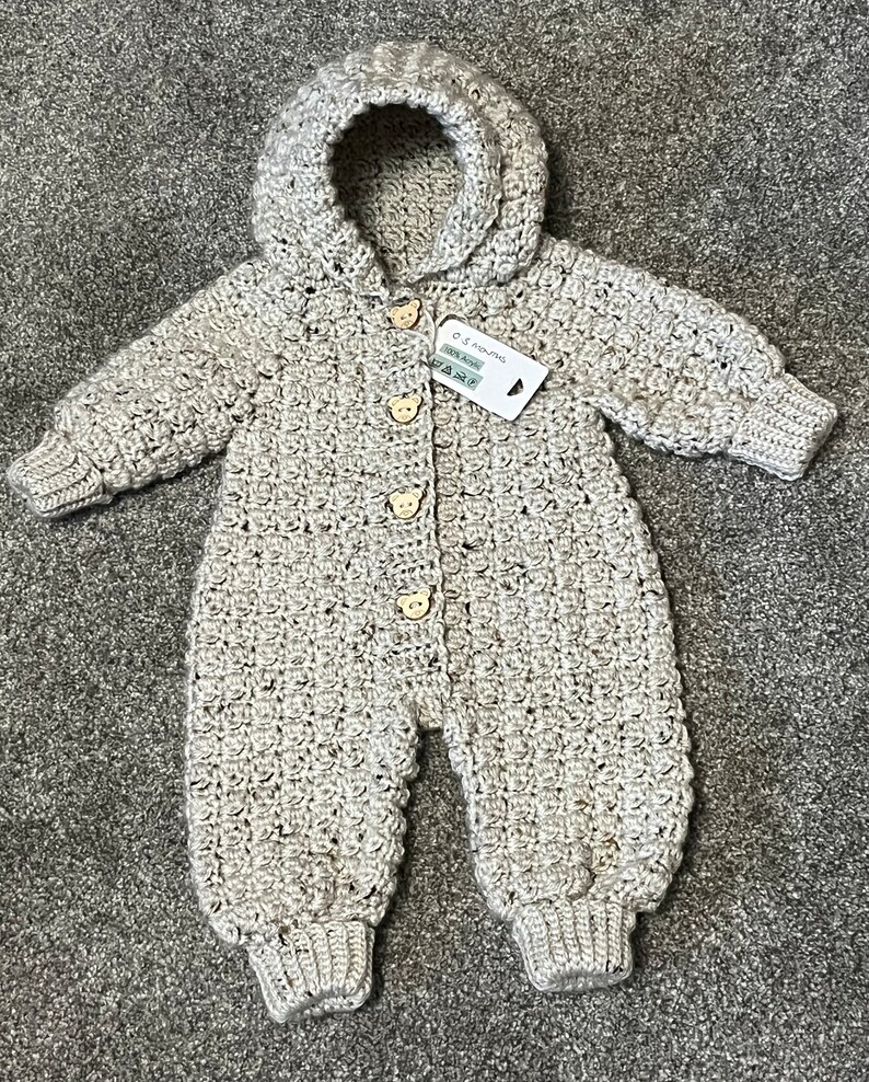 May include: A beige crochet hooded romper featuring long sleeves, ribbed cuffs, and four light brown bear-shaped wooden buttons down the front. A white rectangular tag is attached, displaying the text 0-3 months and 100% Acrylic.