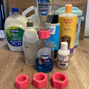 May include: A collection of various cleaning and personal care products, including dish soap, shampoo, and beard wash. Three pink, hexagonal-shaped bottle adapters are in the foreground. The products are arranged on a wooden surface.