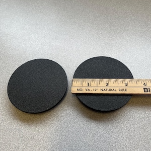 May include: Two black round foam pads, one slightly larger than the other, are placed on a gray surface. A wooden ruler with the text "NO. VA-12" NATURAL RULE" is placed next to the larger pad.